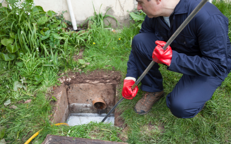 Professional plumber inspecting a drain installation, wearing red gloves and using a tool near an open drainage access point surrounded by grass and plants.